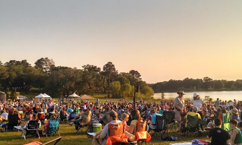 A large outdoor concert crowd gathered by a lake in Southwood at sunset.
